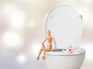 Wooden figurine seating on a toilet and a blood stained toilet paper showing bleeding from hemorhroids