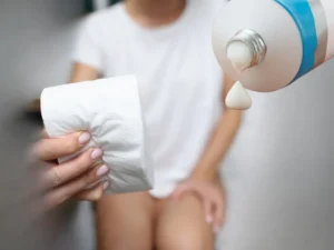 woman seating on a toilet holding toilet paper using to wipe hemorrhoids and a tube of Hydrocortisone cream